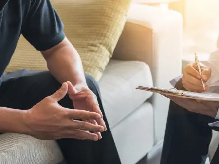 Beratung für Betroffene Zwei Personen im Gespräch auf einem Sofa – eine schreibt auf einem Klemmbrett, die andere gestikuliert. Symbolbild für Beratung bei Glücksspielsucht.